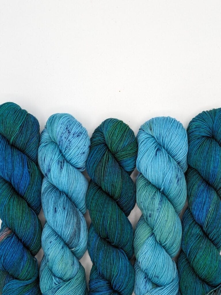 Five skeins of MakerWool yarn in varying shades of blue and green are neatly arranged in a row against a plain white background. The yarn colors range from dark teal to light aqua, with subtle speckles appearing throughout some of the skeins.