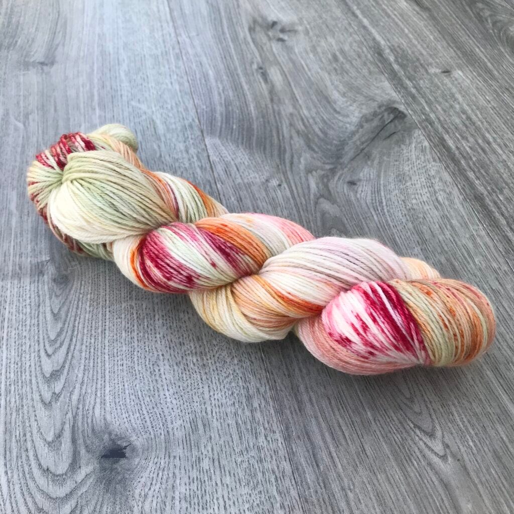 A skein of variegated yarn from Loch Briar Knits lies on a gray wooden surface. The yarn features various hues, including cream, pink, orange, and red, with small speckles of green. The strands are wound together neatly, creating a visually appealing blend of colors and textures.