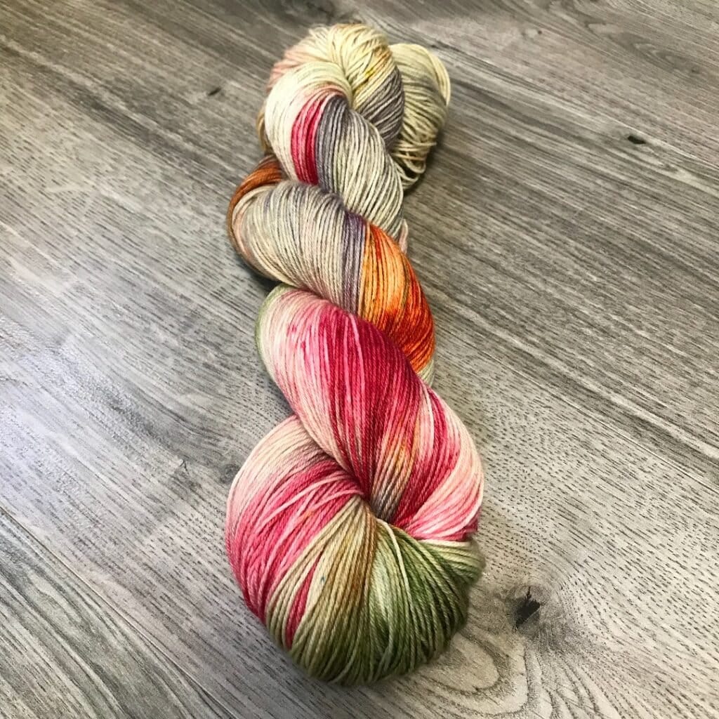 A skein of multicolored yarn lies on a wooden floor. The yarn, from Loch Briar Knits, features a blend of various colors including pink, green, red, orange, beige, and purple twisted together in a visually appealing pattern.