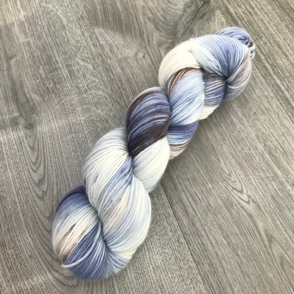A skein of multi-colored Loch Briar yarn is displayed on a wooden surface. The yarn features shades of blue, white, and brown, creating a variegated pattern. The texture appears soft and the yarn is neatly twisted into a hank, ideal for knitting.