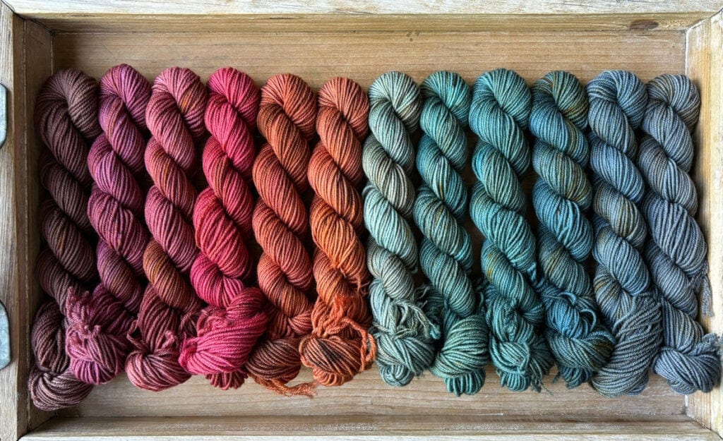 A wooden tray holds tightly arranged skeins of Hidden Pond Yarns in an ombre gradient from deep red through shades of orange, transitioning to green, and ending in blue and grey hues. The eco-friendly, hand-dyed yarns are neatly aligned, showcasing the smooth color progression and exceptional textures.