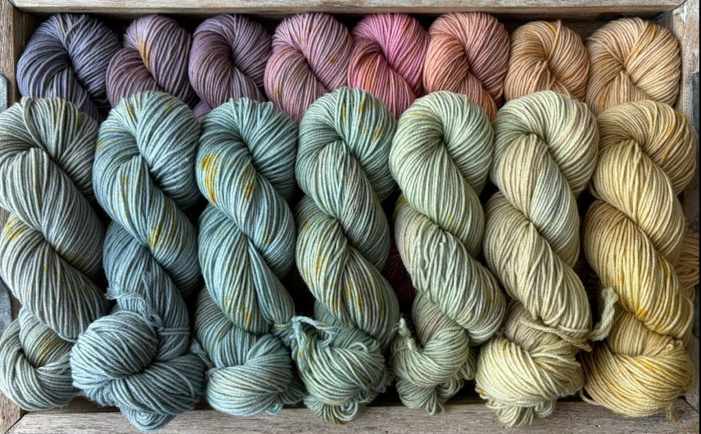 A wooden crate filled with neatly arranged skeins of Hidden Pond Yarns in a gradient of colors, transitioning from purples and mauves on the left, through greens and blues in the middle, to yellows and golds on the right. The yarn is thick and softly textured.