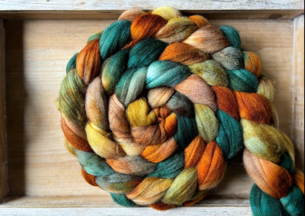 A coiled braid of multicolored wool fibers from Hidden Pond Yarns sits in a wooden tray. The fibers display a rich blend of earthy tones, including green, orange, brown, and yellow.