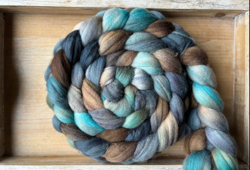 A thick, braided skein of wool roving in shades of teal, blue, green, gray, brown, and beige. The fibers are neatly twisted and coiled inside a wooden tray from Hidden Pond Yarns, creating a visually appealing spiral pattern with natural, earthy tones.