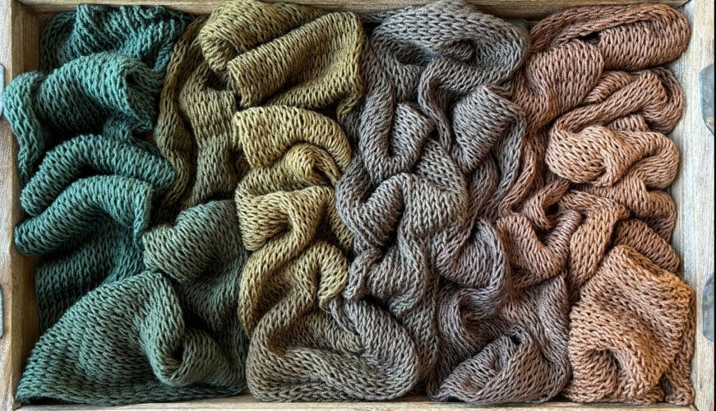 An overhead view of four bundles of knitted fabric in a wooden box, arranged side by side. From left to right, the fabric colors are green, yellow-brown, grey, and reddish-brown. Each bundle from Hidden Pond Yarns is loosely folded, showcasing the texture of the knit pattern.