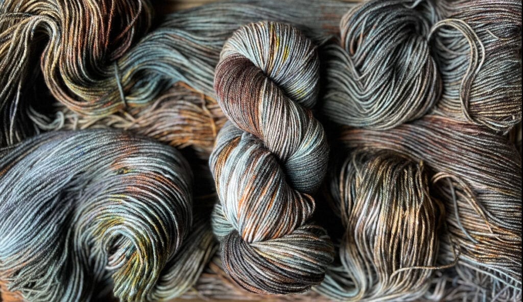 A skein of multicolored yarn from Hidden Pond Yarns, featuring shades of green, brown, and hints of yellow, is neatly wound in the center. Surrounding it are loose, unwound strands of the same yarn, creating a textured and visually rich backdrop.