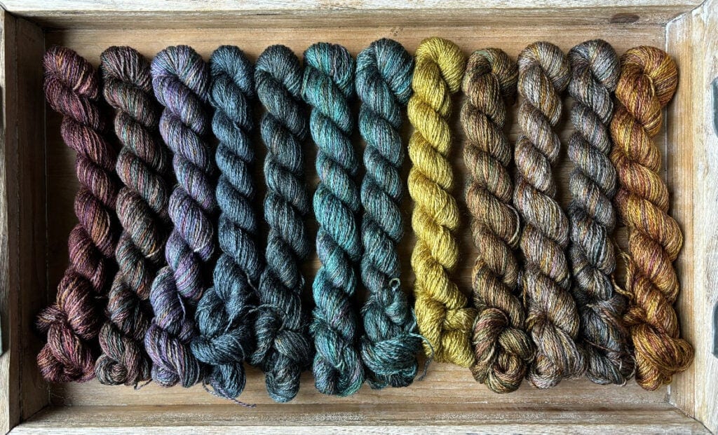 A wooden tray holds twelve skeins of Hidden Pond Yarns neatly arranged in a row. The yarn colors transition seamlessly from deep purples and blues on the left, through greens and teals in the middle, to yellows and browns on the right.