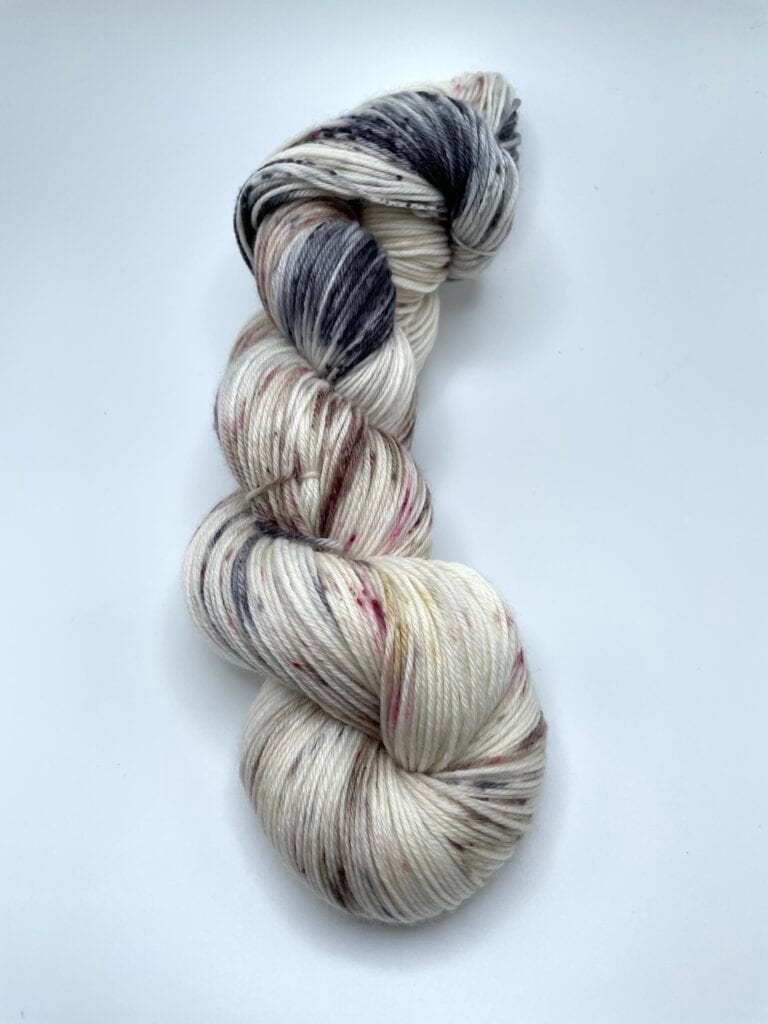 A twisted skein of yarn in a mix of white, black, and light pink hues is showcased against a plain white background. The yarn, crafted by Loch Briar Knits, is neatly wound, displaying its multicolored strands intertwined in an artistic manner.
