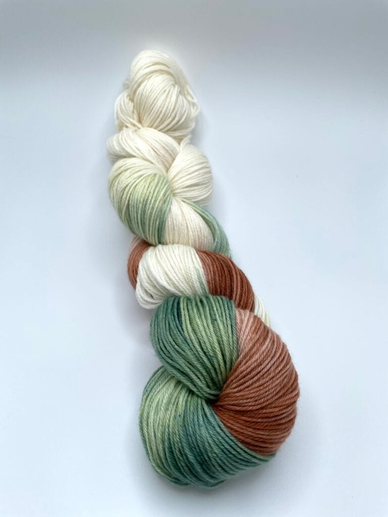 A twisted skein of yarn from Loch Briar Knits features a blend of colors including white, cream, green, and brown. The yarn has a smooth texture and is neatly coiled against a plain white background.