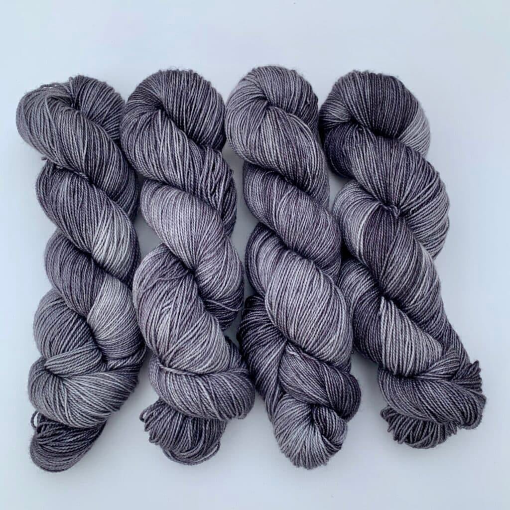 Four skeins of Loch Briar grey yarn are neatly arranged side by side against a white background. The yarn features a mix of light and dark grey tones, giving it a speckled appearance—perfect for your next Knits project.