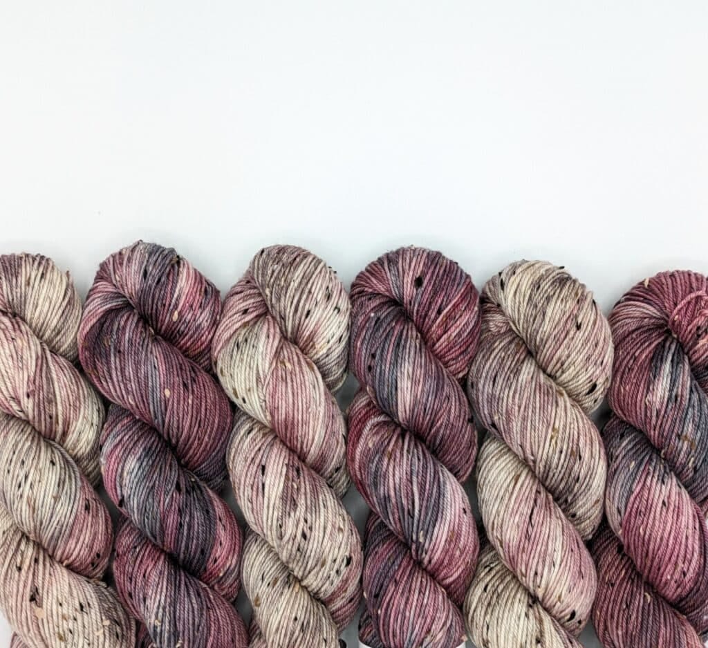Five skeins of MakerWool yarn are arranged in a row against a plain white background. The yarn is variegated with shades of pink, purple, gray, and cream, and has a speckled pattern with dark spots throughout.