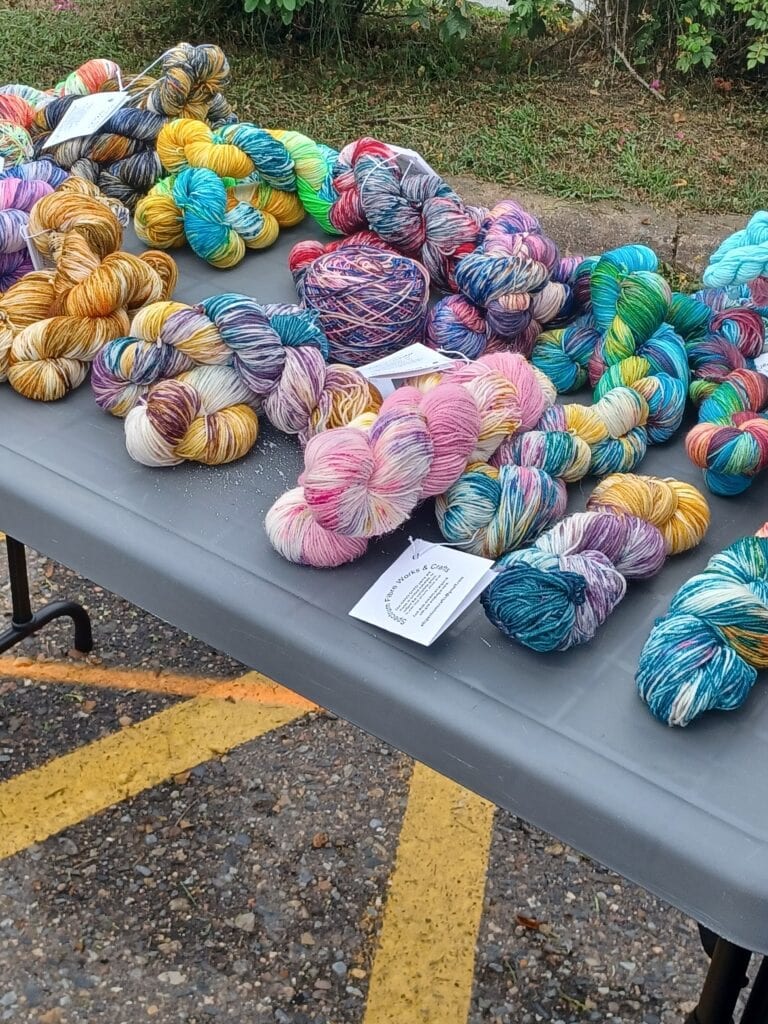 A table displays an assortment of colorful yarn skeins in various patterns, including pink, yellow, blue, and multicolored. Among the vibrant fibers and scattered business cards lies an air of creativity. The background showcases some greenery and a yellow-painted asphalt space, blending nature's beauty with an auto draft of ideas.