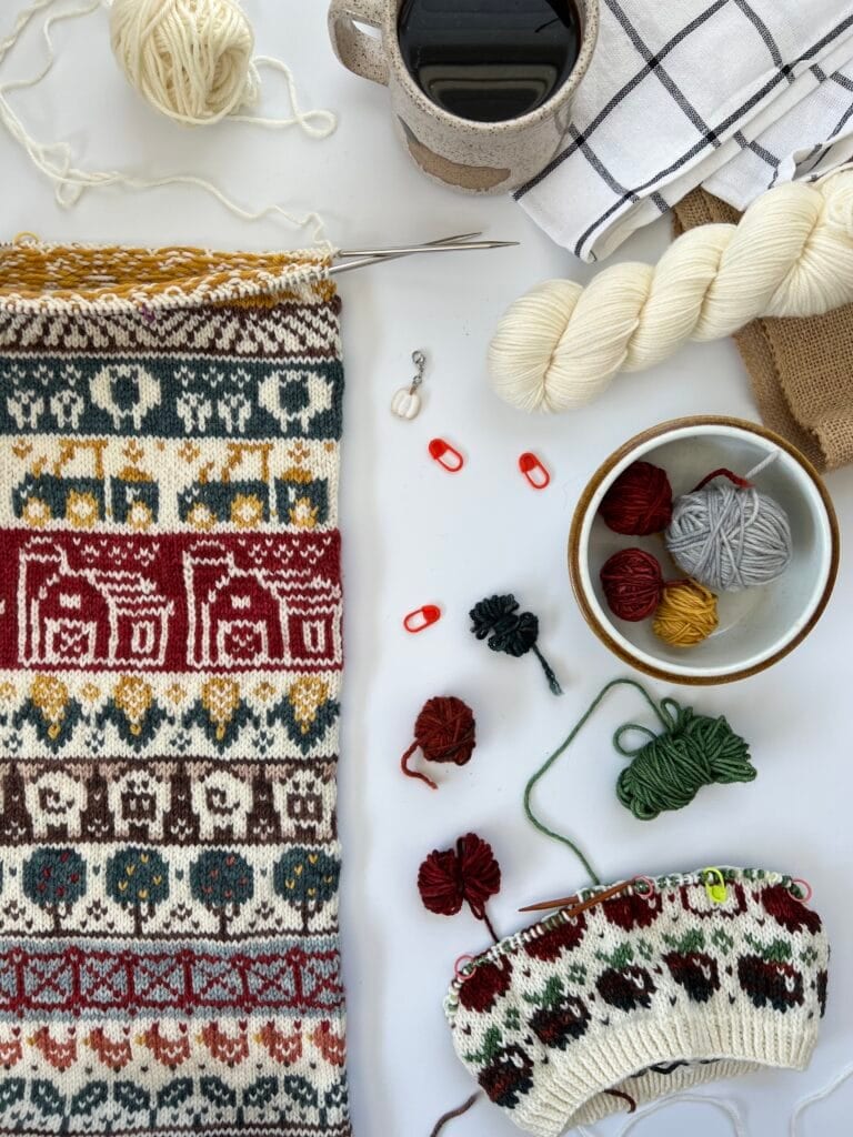 A knitting project in progress showcasing intricate patterns in various colors, surrounded by knitting needles, yarn from Baa-Realis Fibre, stitch markers, a coffee cup, and a checkered cloth on a white surface.