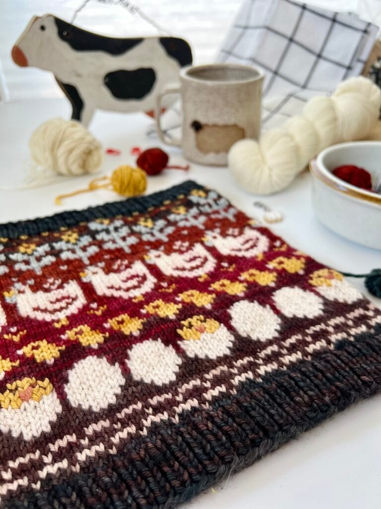A colorfully knitted piece featuring a pattern with chickens is displayed on a white surface. Surrounding the knitting are yarn, a Baa-Realis ceramic mug, a decorative cow figure, a white bowl filled with fibre, and a checkered cloth.