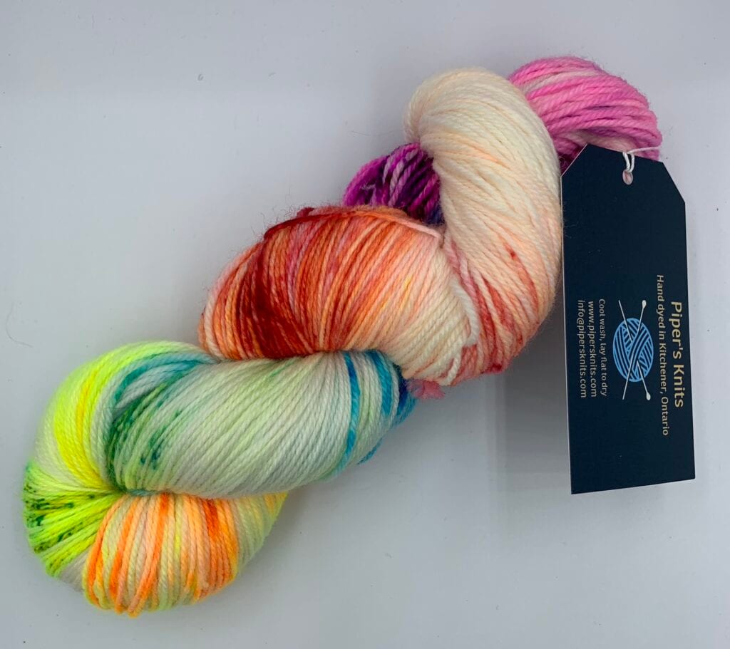 A skein of multicolored yarn with hues of pink, purple, orange, yellow, green, and blue is twisted together with a black tag attached. The tag prominently features 