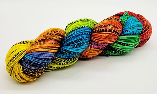 A colorful skein of yarn lies against a plain white background. The vibrant array of colors—red, orange, yellow, green, blue, and purple—with black stripes interspersed throughout is perfect for all those Nerds with Needles crafting up their next masterpiece.