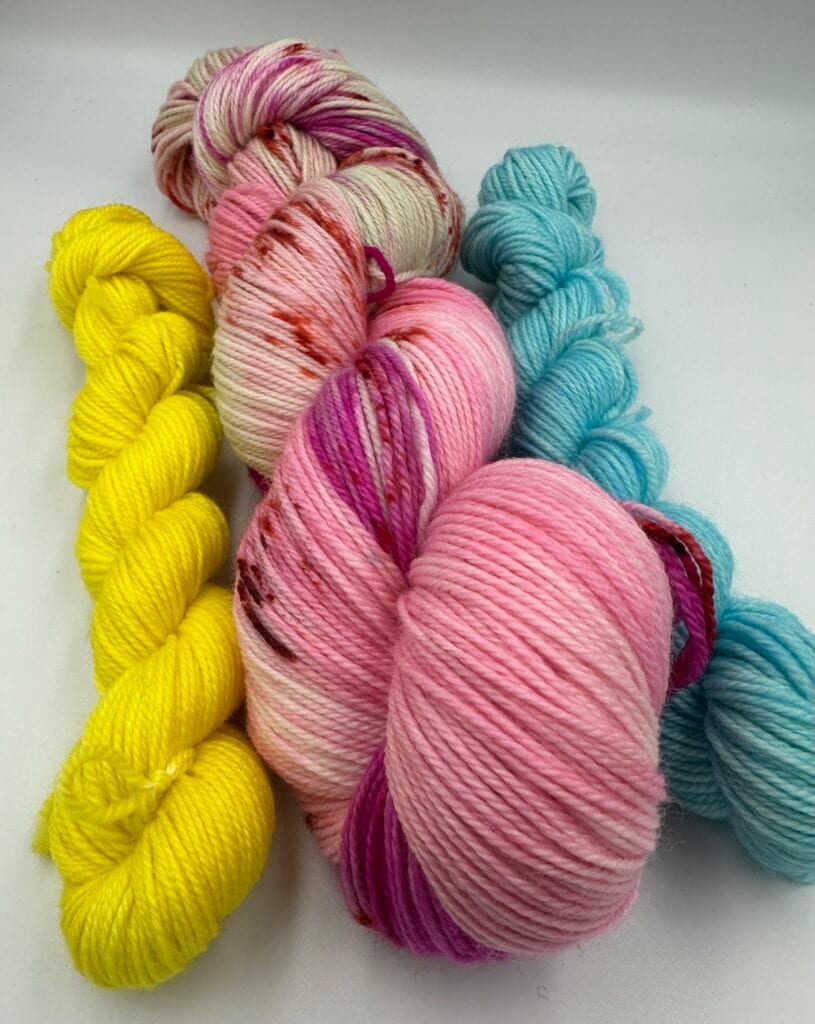 Four skeins of Piper's Knits yarn are arranged on a white background. The colors are bright yellow, pink with splashes of red and purple, solid light pink, and turquoise blue. The yarn appears soft and plush, indicating a likely wool or wool blend.