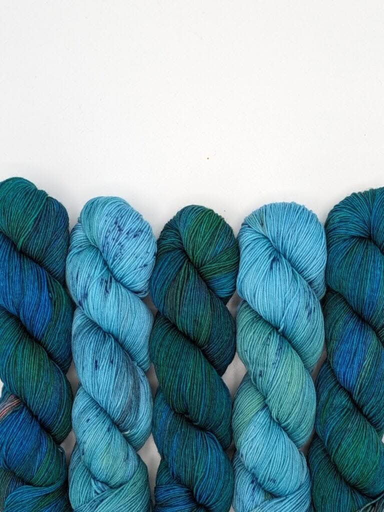 Five skeins of MakerWool yarn in varying shades of blue and green are neatly arranged in a row against a plain white background. The yarn colors range from dark teal to light aqua, with subtle speckles appearing throughout some of the skeins.