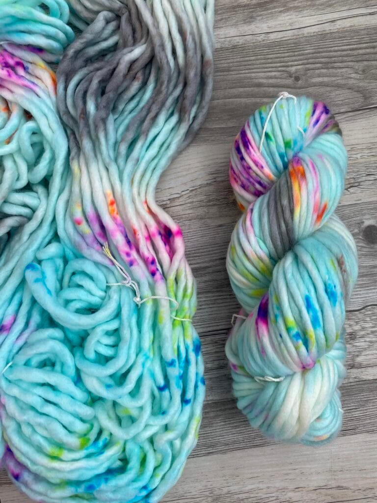 A skein and a bundle of thick, multicolored yarn lie on a wooden surface. The vibrant Nerdy Knits yarn showcases a base of light blue with splashes of pink, yellow, green, and purple. The colors pop against the soft, chunky texture of this delightful crafting material.