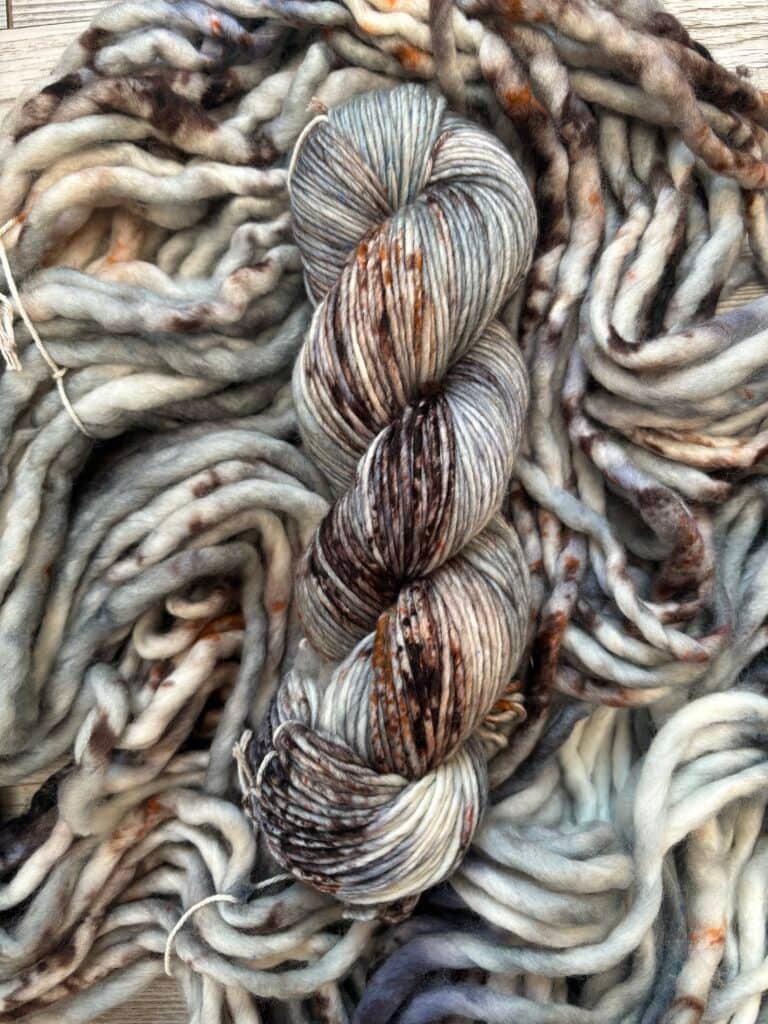 Close-up of a skein of variegated yarn placed on a backdrop of twisted, thick fibers. The Nerdy Knits yarn features a mix of colors, including shades of gray, brown, and white with occasional specks of rust. The texture appears soft and chunky, suitable for cozy projects.