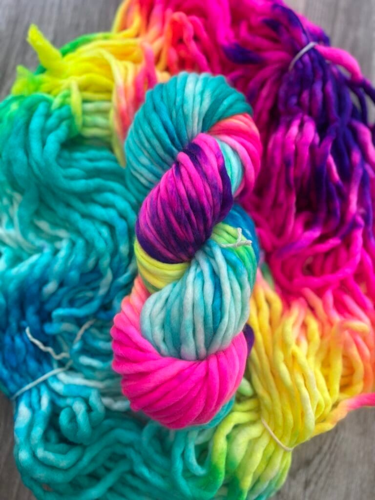 A colorful skein of yarn in vibrant shades of pink, blue, yellow, green, and purple is displayed on a wooden surface. The yarn is slightly unraveled around the skein, revealing its soft and thick texture. This Nerdy Knits creation showcases a burst of hues perfect for any knitting enthusiast.