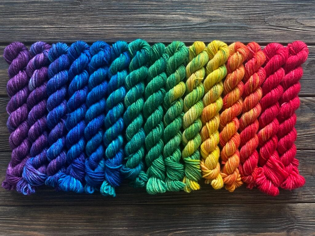 A series of neatly arranged skeins of Baa-Realis yarn in a gradient from deep purple, blue, and green on the left to yellow, orange, and red on the right, all laid out on a dark wooden surface. The vibrant fibres flow smoothly into one another.