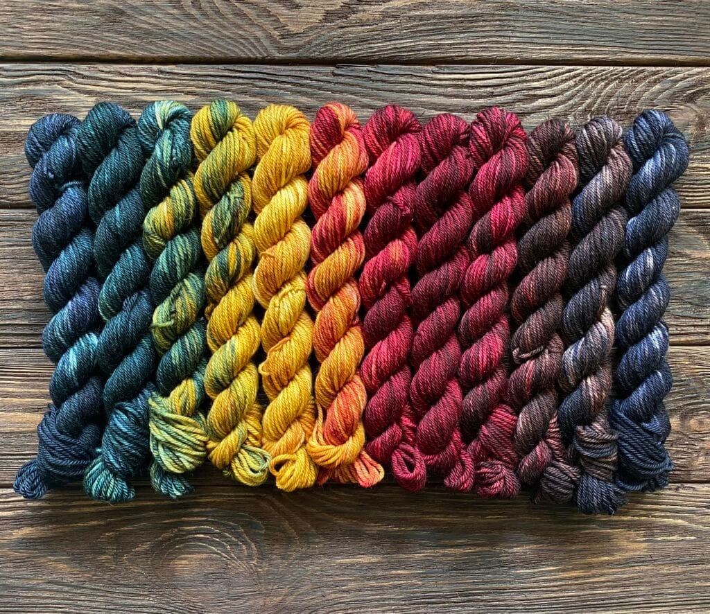 Several skeins of Baa-Realis yarn are neatly lined up in a rectangular formation on a wooden surface. The vibrant fibre colors transition smoothly from dark green on the left through blue, yellow, orange, red, and brown, creating a mesmerizing gradient effect.