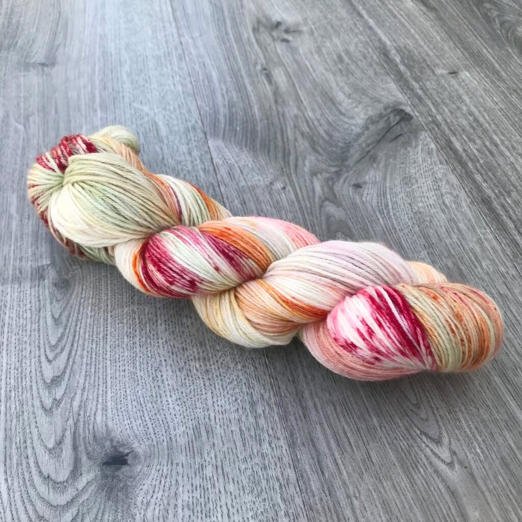 A skein of variegated yarn from Loch Briar Knits lies on a gray wooden surface. The yarn features various hues, including cream, pink, orange, and red, with small speckles of green. The strands are wound together neatly, creating a visually appealing blend of colors and textures.