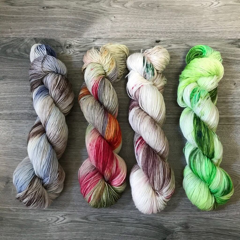 Four skeins of multicolored yarn from Loch Briar Knits are arranged side by side on a gray wooden surface. The yarn colors range from earthy tones and grays with mixed speckles to vibrant greens and reds. Each skein has a unique color pattern, showcasing a variety of dye techniques.