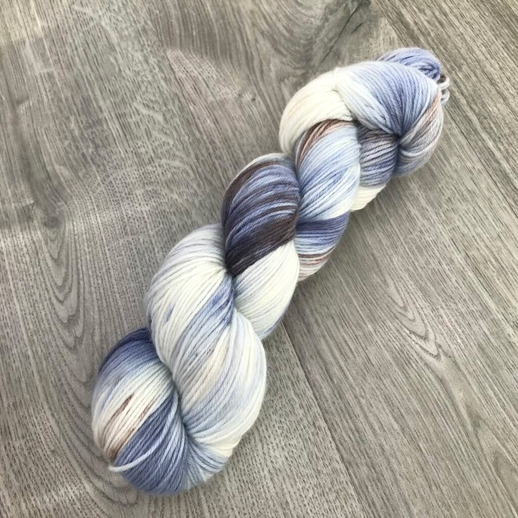 A skein of multi-colored Loch Briar yarn is displayed on a wooden surface. The yarn features shades of blue, white, and brown, creating a variegated pattern. The texture appears soft and the yarn is neatly twisted into a hank, ideal for knitting.