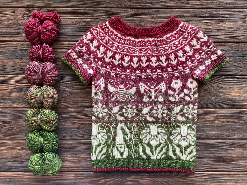 A hand-knitted short-sleeve sweater with intricate maroon, white, and green patterns is displayed on a wooden surface. Yarns in matching maroon and green hues are placed in balls beside the Baa-Realis sweater on the left, showcasing the high-quality fibre used.