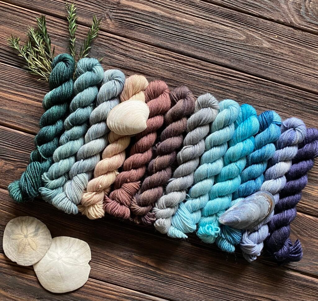 A collection of variously colored yarn skeins arranged in rows on a rustic wooden surface. The yarn colors transition from shades of green to blue to purple. Interspersed with seashells and sprigs of greenery, this Baa-Realis display beautifully enhances the natural theme with exquisite fibre artistry.