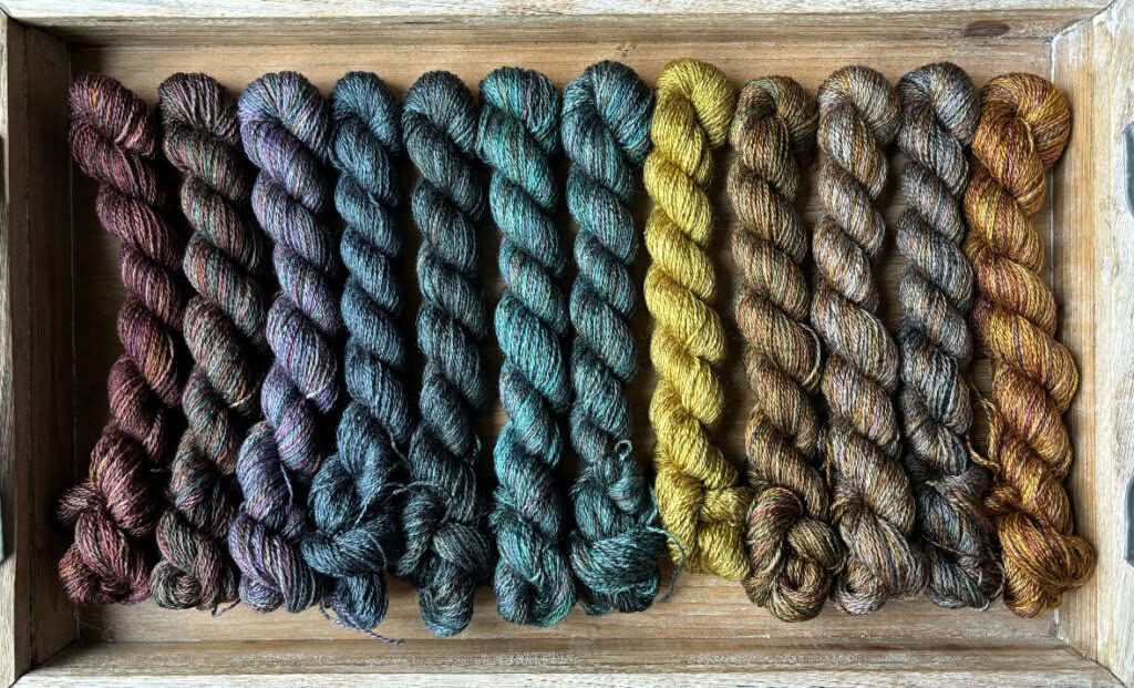 A wooden tray holds twelve skeins of Hidden Pond Yarns neatly arranged in a row. The yarn colors transition seamlessly from deep purples and blues on the left, through greens and teals in the middle, to yellows and browns on the right.
