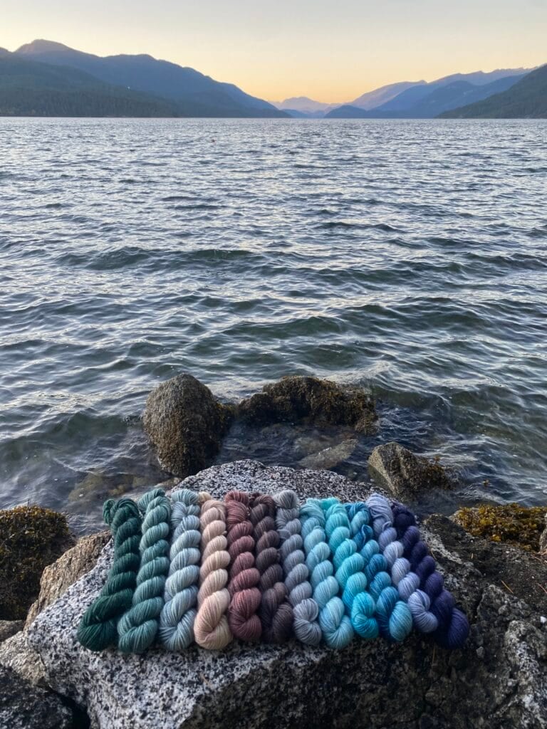A collection of Baa-Realis Fibre yarn skeins in various earth and ocean hues is neatly arranged on a grey, speckled rock. The rock sits by the shore of a calm, expansive lake, surrounded by distant mountains under a clear, soft-lit sky. The scenery evokes a peaceful natural atmosphere.