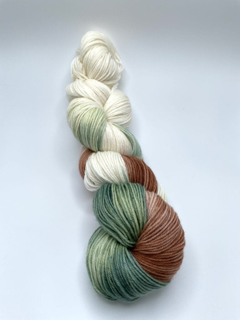 A twisted skein of yarn from Loch Briar Knits features a blend of colors including white, cream, green, and brown. The yarn has a smooth texture and is neatly coiled against a plain white background.