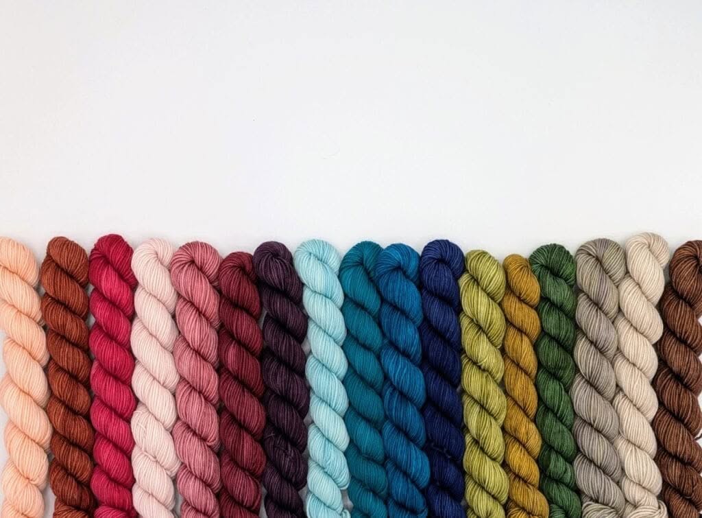 A neatly arranged row of colorful MakerWool yarn skeins is shown against a plain white background. The skeins display a gradient from pinks and reds on the left, transitioning through blues and greens, and ending with neutrals on the right.