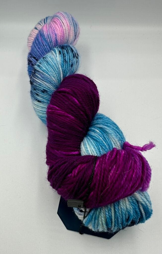 A twisted skein of yarn displaying a vibrant gradient of colors, including shades of pink, purple, blue, and white. The yarn has a soft and thick texture, with small speckles of black scattered throughout. Set against a plain light gray background, it looks like an artist's auto draft brought to life.