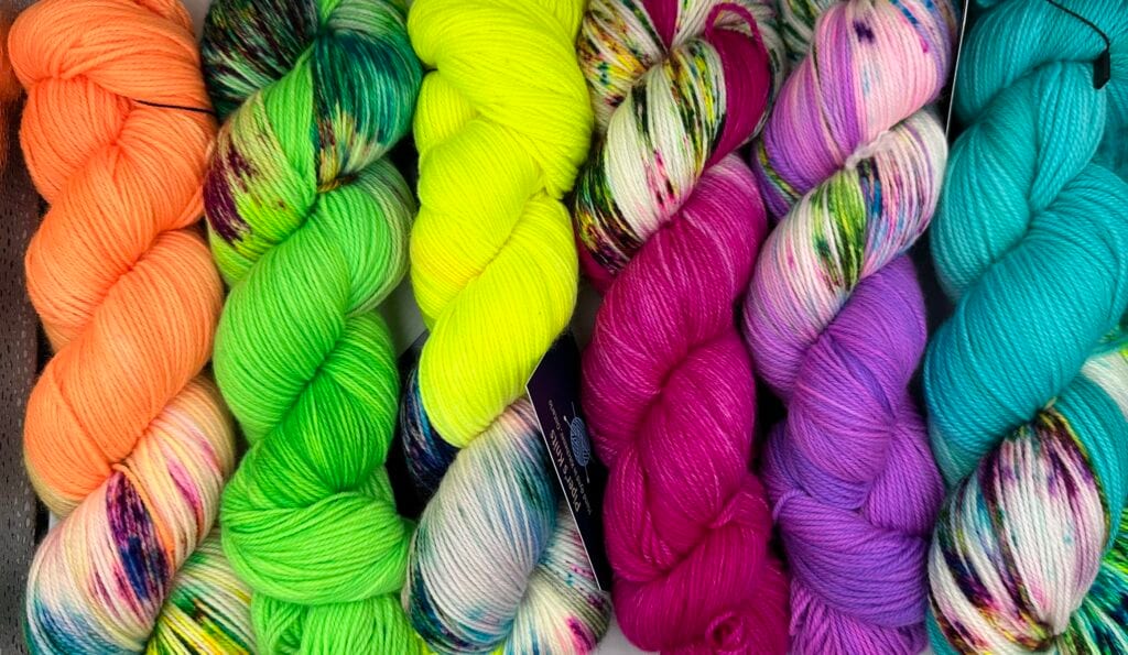 A vibrant assortment of skeins of yarn is displayed in a row at Piper's Knits. From left to right, the colors include peach, green, yellow, multi-colored white and green, fuchsia, purple, and blue with multicolored specks. Each skein has a distinct, bright hue and texture.