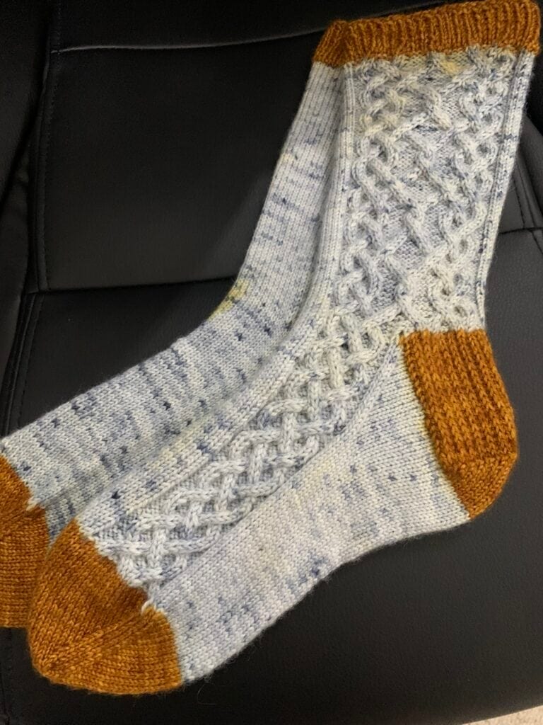 A pair of hand-knitted socks from Piper's Knits featuring intricate cable patterns, predominantly light gray with flecks of darker color. The toe, heel, and cuff areas are knitted with contrasting brown yarn. The socks are placed on a black leather surface.